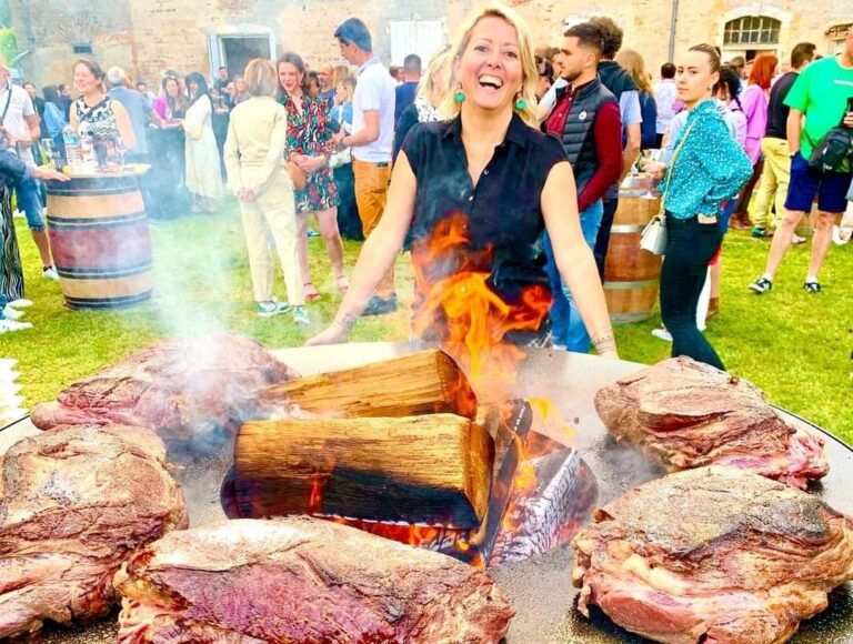 Cuisinière rieuse devant un braséro de 100 chargé de viandes et de bûches