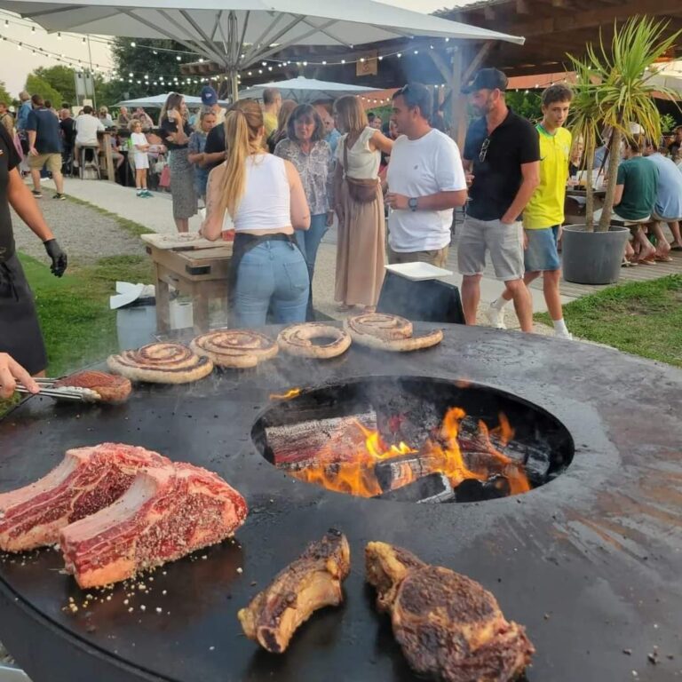 Public devant un braséro rond de 150 en évènement : côtes de bœuf, saucisses en couronne et flammes