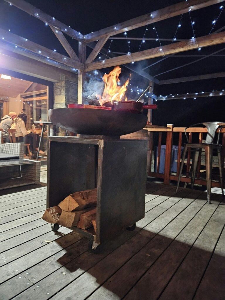Braséro rond sur meuble avec réserve de bûches, flammes sur la terrasse éclairée d'un restaurant, la nuit