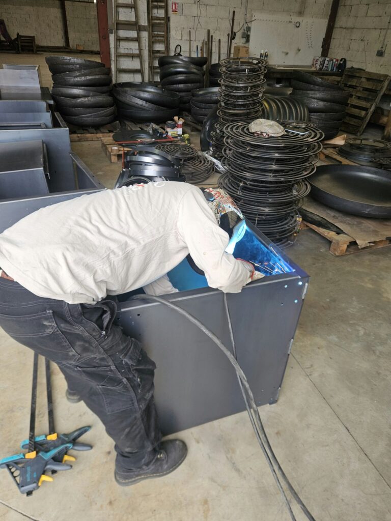 Dans l'atelier, un soudeur vu de dos, casque baissé, penché sur un caisson en tôle qu'il soude ; derrière lui, des bols et des grilles de braséros empilés