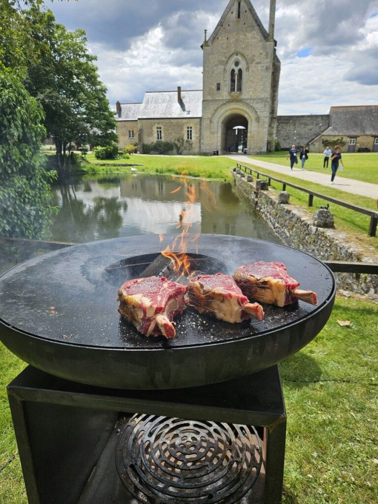 Braséro rond près d'un étang, abbaye en arrière-plan, viandes sur la plancha