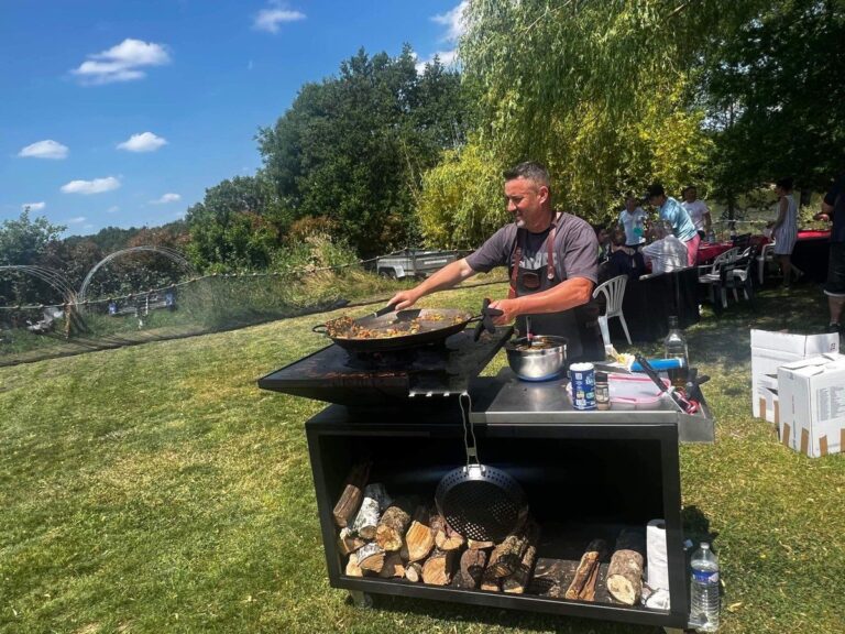 Cuisinier à la poêle devant un braséro carré sur meuble noir avec réserve de bûches, au jardin