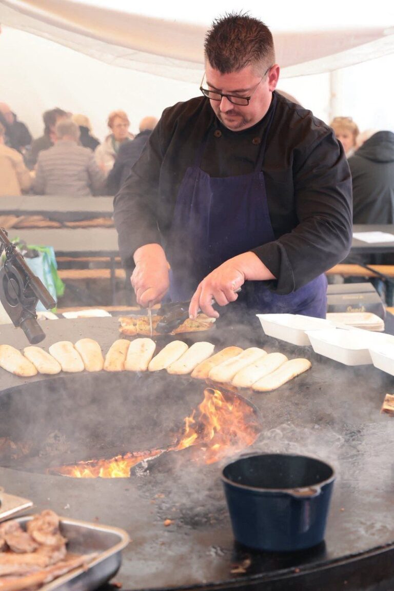 Cuisinier en tablier bleu devant un braséro avec une marmite, public attablé au fond