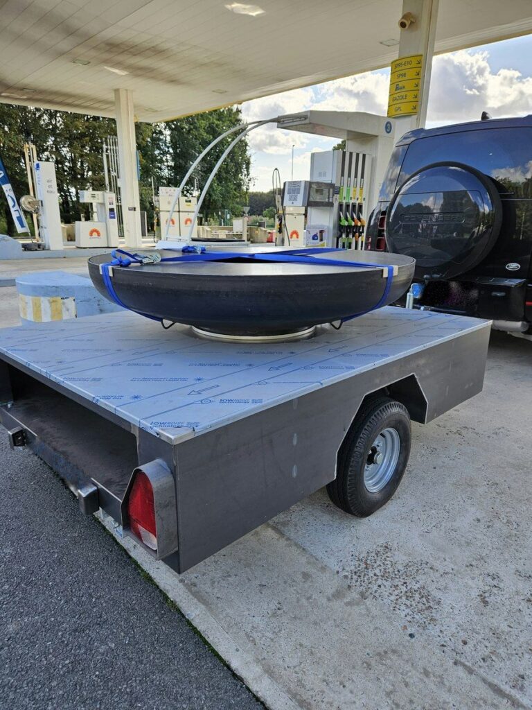 Braséro rond sanglé sur sa remorque à plateau en inox, vue de trois quarts arrière