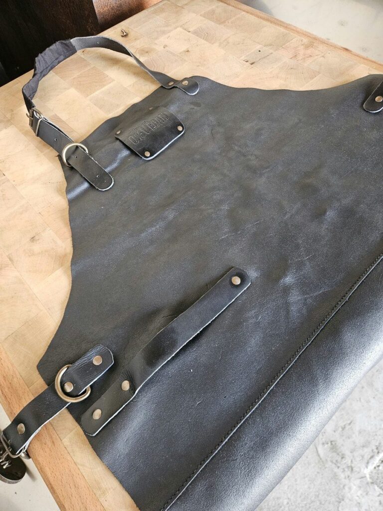 Tablier en cuir noir vu en biais sur un billot de boucher, avec sa bretelle réglable et ses attaches à anneau